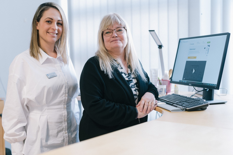 Två kvinnor står vid sidan om ett skrivbord med datorskärm.