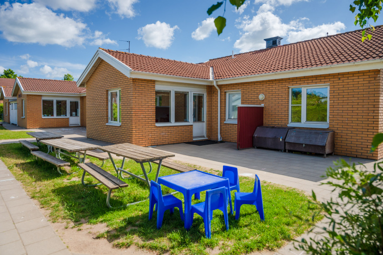 Enplans hus i vinkel, orange tegel och tak. I förgrunden står sittgrupper på en liten gräsyta