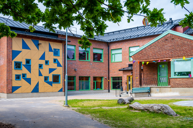 Rött tegelhus i två plan. Byggt i vinkel. Den ena huskroppen är förlängd med en gulputsad del med blå dekorationer. I förgrunden asfalt, gräs och en trädgren med gröna löv.