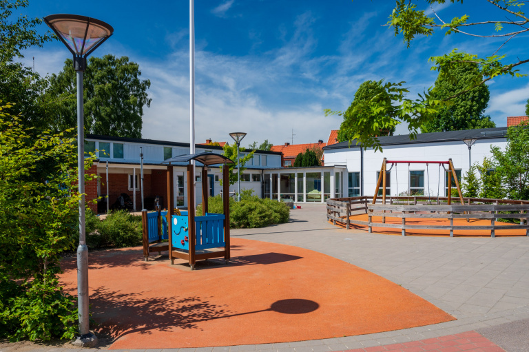I förgrunden lekplats i bakgrunden vita skolbyggnader med platta tak.
