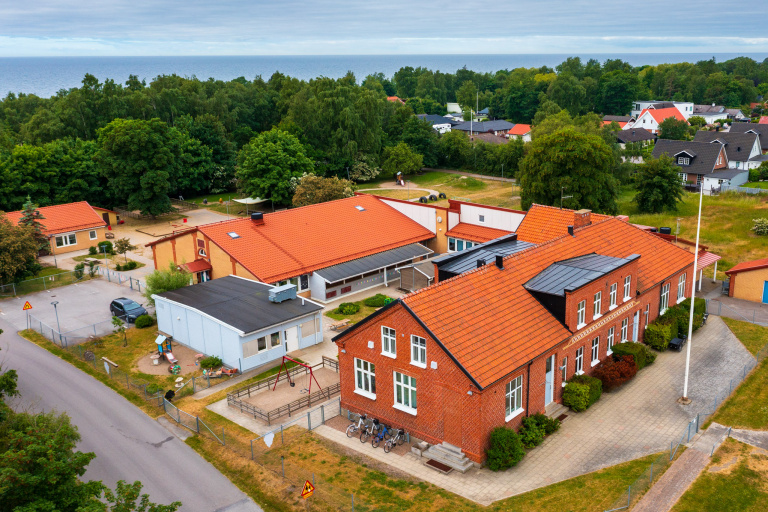 Bild tagen från ovan. Större röd en och en halvplans tegelhus i äldre modell, samt en större gul byggnad i ett plan med rött tak. I bild syns även ett mindre vitt hus med svart tak och omgivande mark som är indelat i områden av gräs och grus