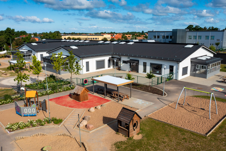 Vit, modern enplansbyggnad i vinkel med svart tak. Delar av lekplats och stenlagd utemiljö syns i bilden, som är fotograferad ovanifrån.