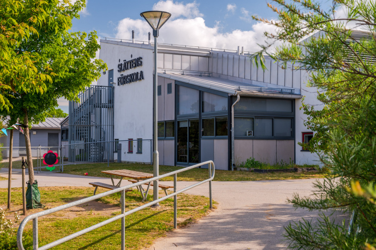 I förgrunden lyktstolpe, gräsmatta och bänk. I bakgrunden vit, modern byggnad med pulpettak och texten "Slättens förskola"