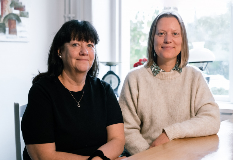 Två leende kvinnor sitter vid ett bord och tittar in i kameran.