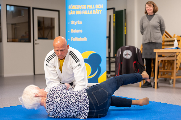 Man i judodräkt övar fall med kvinna på mjuk matta.