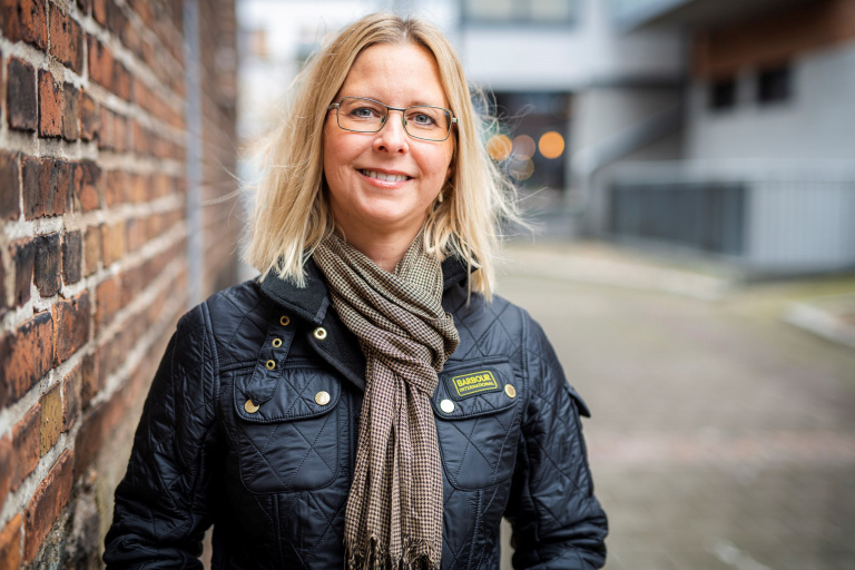 Kvinna står vid tegelvägg och tittar leende in i kameran.