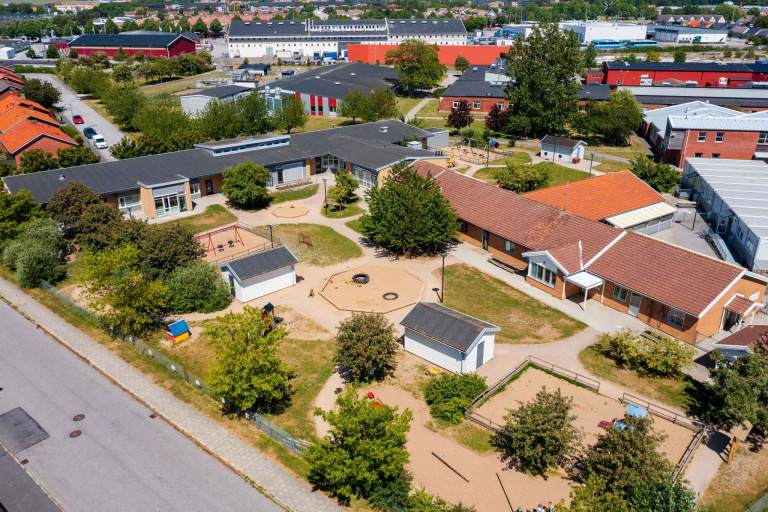 Bild tagen ovanifrån. I bilden syns en avlång byggnad med rött tak samt två mindre vita huse med svart tak. Husen ligger på en skolgård med grus, grönytor, träd och buskar.