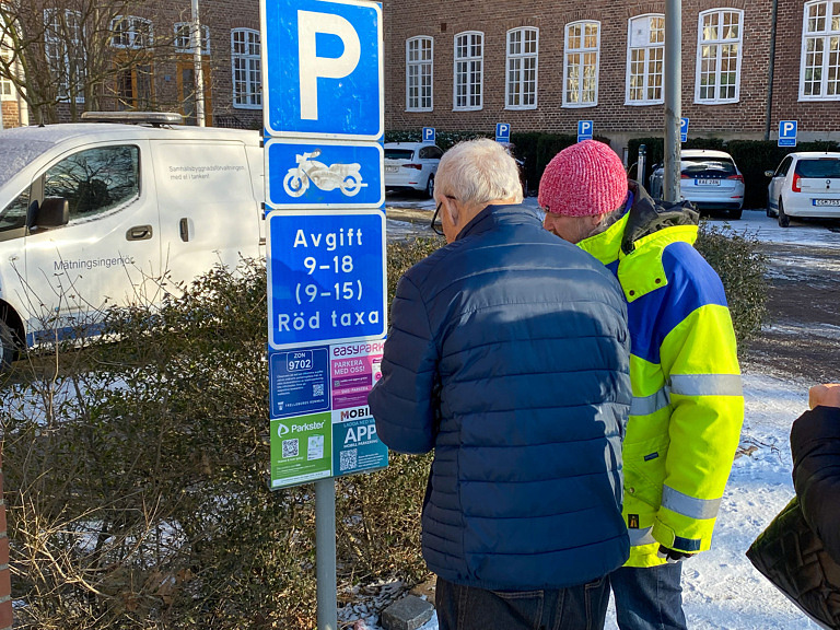 Man i blå jacka och man i gul jacka vid P-skylt