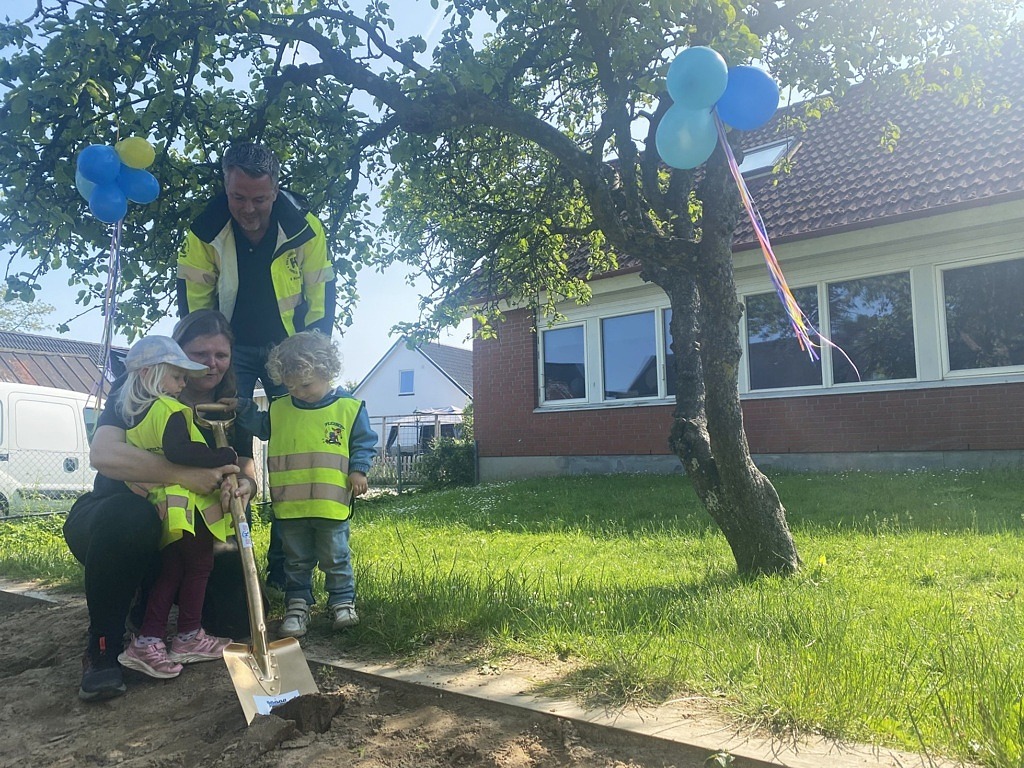 två barn får hjälp av två vuxna att gräva med en stor guldig spade