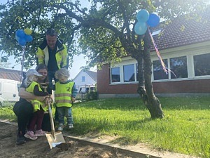 två barn får hjälp av två vuxna att gräva med en stor guldig spade
